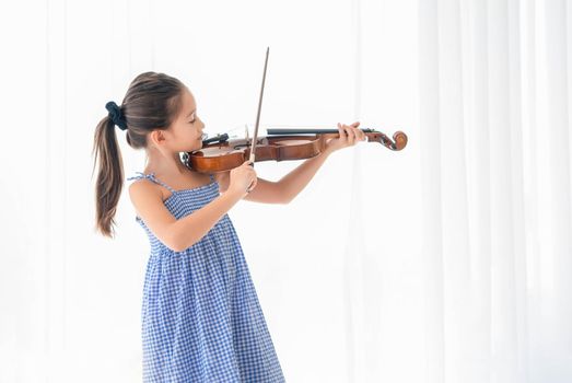 Cute little girl playing violin in bedroom with white curtain background. Musical and people lifestyle. Education and recreation concept. Asian musician violinist kids doing hobby and leisure at home.