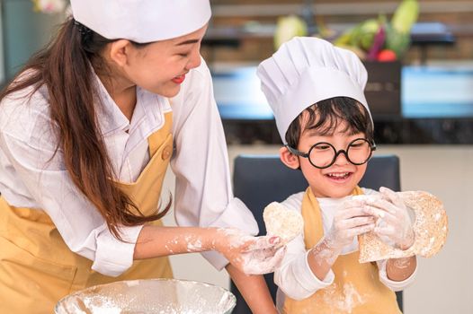 Beautiful woman and cute little Asian boy with eyeglasses, chef hat and apron playing and baking bakery in home kitchen funny. Homemade food and bread. Education and learning concept. Thai person