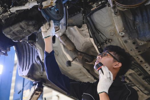 Asian male mechanical hold and shining flashlight to examine car under chassis of automotive vehicle. Safety suspension inspection check service maintenance for customer before road trip concept