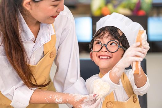 Beautiful woman and cute little Asian boy with eyeglasses, chef hat and apron playing and baking bakery in home kitchen funny. Homemade food and bread. Education and learning concept. Thai person