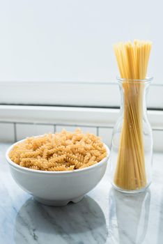 White bowl of spiral macaroni and tied spaghetti stand erect at windows of kitchen room. Food and ingredients concept. Italian food theme