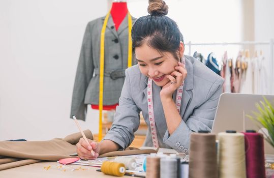 Attractive Asian female fashion designer  working in home office workshop. Stylish fashionista woman creating new cloth design collection. Tailoring and sewing. People lifestyle and occupation concept