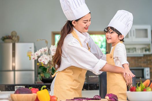 Happy beautiful Asian woman dress up cute little boy chef outfit for prepare to cooking in home kitchen. People lifestyles and Family. Homemade food and ingredients concept. Two Thai people life