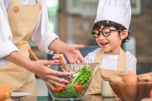 Cute little Asian happy boy interested in cooking with mother funny in home kitchen. People lifestyles and Family. Homemade food and ingredients concept. Two Thai people making ketogenic diet salad