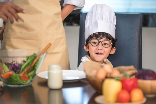 Closeup of Asian cute little boy wearing chef hat and apron with mother in home kitchen. Thai people and lifestyles concept. Job occupation and Education learning concept. Happy family time theme