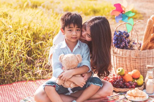 Little Asian boy kissed at cheek by his mom in meadow when doing picnic. Mother and son playing together. Celebrating in Mother day and appreciating concept. Summer people and lifestyle theme.