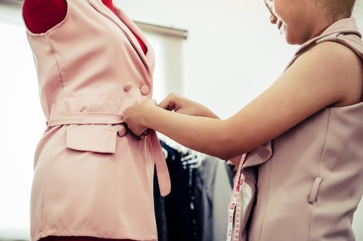 Asian female fashion designer girl making fit on the formal dress uniform clothes on mannequin model. Fashion designer stylish showroom. Sewing and tailor concept. Creative dressmaker stylist.
