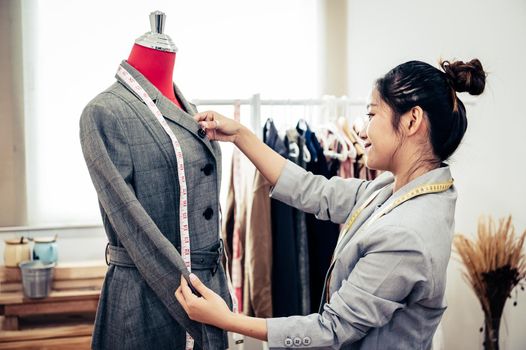 Asian female fashion designer girl making fit on the formal suit uniform clothes on mannequin model. Fashion designer stylish showroom. Sewing and tailor concept. Creative dressmaker stylist.