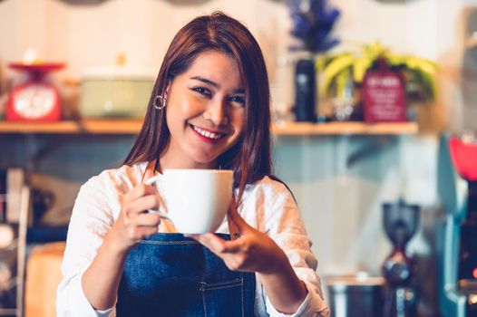 Asian female barista making cup of coffee. Young woman holding white coffee cup while standing behind cafe counter bar in restaurant background. People lifestyles and Business occupation concept.