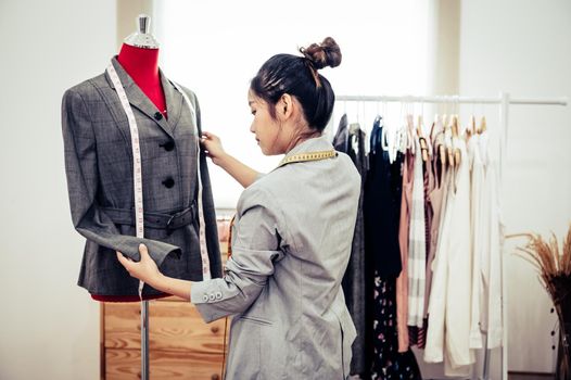 Asian female fashion designer girl making fit on the formal suit uniform clothes on mannequin model. Fashion designer stylish showroom. Sewing and tailor concept. Creative dressmaker stylist.