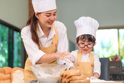 Beautiful woman and cute little Asian boy with eyeglasses, chef hat and apron playing and baking bakery in home kitchen funny. Homemade food and bread. Education and learning concept. Thai person