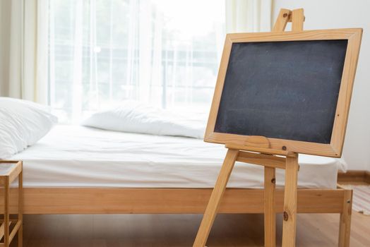 Black chalkboard on easel stand in bedroom with white soft bed background. Interior and decoration concept.