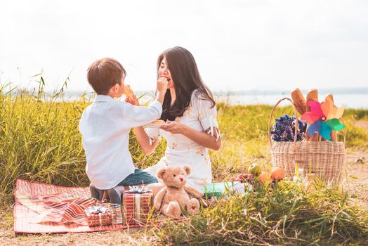 Little Asian boy his mom feeding snack each other in meadow when doing picnic. Mother and son playing together. Celebrating in Mother day and appreciating concept. Summer people and lifestyle theme.