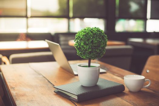Small decorate tree, laptop, notebook and coffee cup in on wooden table in coffee shop. Relaxation and Business Co-working space concept