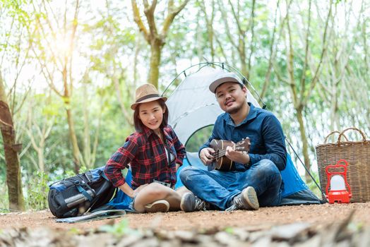 Asian couple camping in the forest. Man playing ukulele with woman in front of camping tent. Lamp and basket and backpack element. People and outdoor lifestyles concept. Adventure and travel theme.