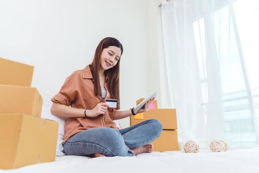 Beauty Asian woman using tablet and credit card. Start up small business entrepreneur SME and checking order list in bedroom, Young happy freelance woman shopping online marketing or sending parcel