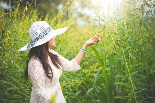 Beauty Asian woman in white dress and wing hat walking in rapeseed flower field background. Relaxation and travel concept. Wellness happy life of girl on vacation. Nature and people lifestyle