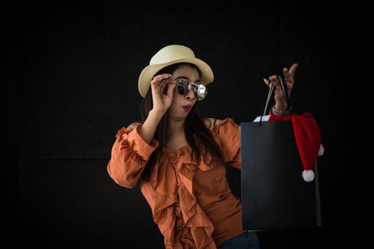 Asian shopping woman surprised with Black Friday shopping bag and Santa Claus hat inside on black background. Shopaholics and beauty fashion theme.