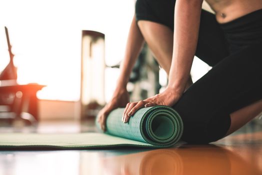Close up of sporty woman folding yoga mattress in sport fitness gym training center background. Exercise mat rolling keeping after yoga class. Workout and sport training concept. Hands on carpet