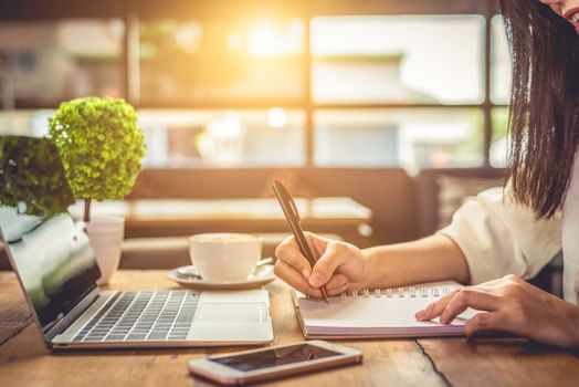 Close up of woman hand writing business project planning in notebook with laptop and smart phone at coffee shop. People and technology concept. Freelance and Lifestyle theme. Entrepreneur and working