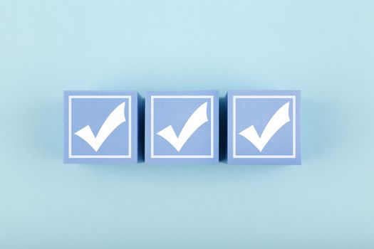Three white checkmarks on toy cubes in a row on bright pastel blue background. Concept of questionary, kids related checklist, to do list, planning, business or verification.