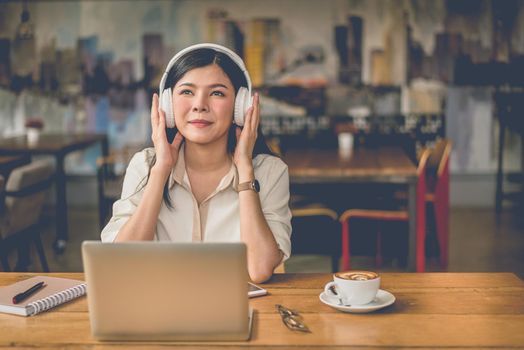 Happy Asian woman relaxing and listening music in coffee shop with computer laptop and coffee cup. People and lifestyles concept. Freelance and outdoors workplace outdoors theme.