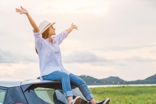Happy Asian woman spread arms on car roof under sunset at seaside with nature  background during travel in holiday. Confidence cheerful girl with dawn sky. Sun sea and mountain. Vacation concept