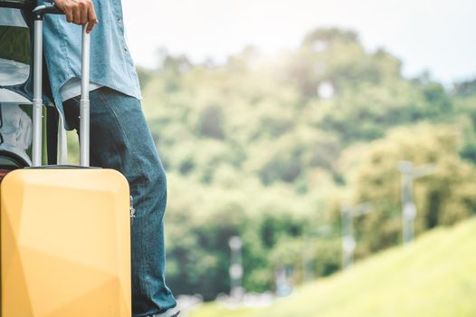 Closeup of man legs resting during travel on road trip with yellow suitcase on summer nature background. Transportation and people lifestyles concept. Tourist standing outdoors on vacation tour