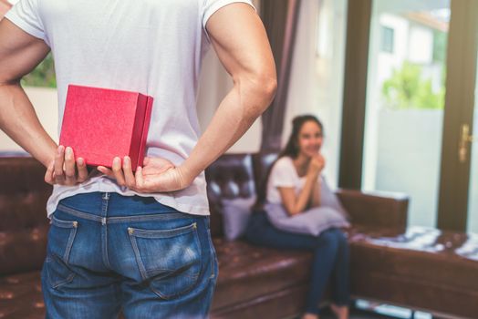 Guy holding gift behind him for surprise his waiting girlfriend at their home. Selective focus on gift.