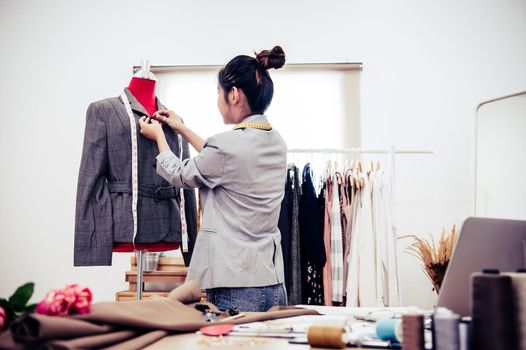 Asian female fashion designer girl making fit on the formal suit uniform clothes on mannequin model. Fashion designer stylish showroom. Sewing and tailor concept. Creative dressmaker stylist.