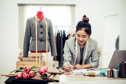 Attractive Asian female fashion designer  working in home office workshop. Stylish fashionista woman creating new cloth design collection. Tailor and sewing. People lifestyle and occupation concept