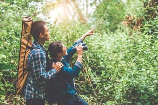 Couple of Asian people holding binoculars telescope in forest looking forward to destination. People lifestyles and leisure activity. Nature and backpacker traveling jungle background. Bird watching