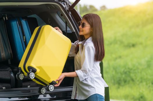 Asian woman lifting yellow suitcase into SUV car during travel in long weekend trip. People lifestyles and transportation concept. Girl put luggage from car trunk to camping. Selective focus on car