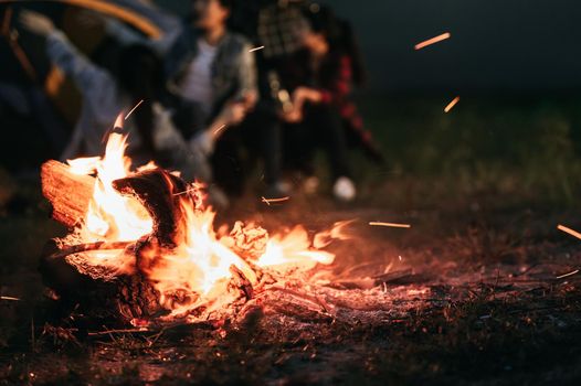 Sparking bonfire with tourist people sit around bright bonfire near camping tent in forest in summer night background. Group of student at outdoor fire fuel. Travel  activity and long vacation weekend