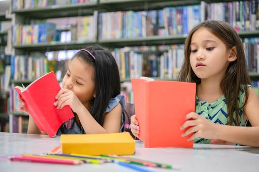 Two little cute girls multi-ethnic friends reading books together in school library. People lifestyles and Education learning concept. Happy friendship kids doing leisure activity for examination test