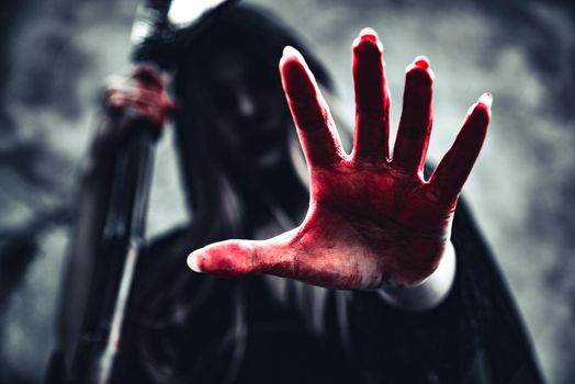 Witch showing bloody hand with reaper. Female demon angel in black clothes and hood on grunge wall background. Halloween day and Mystery concept. Fantasy of magic theme. Afterlife and Death concept.