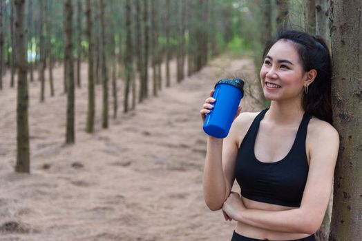 Beauty Asian sport woman resting and holding drinking water bottle and relaxing in middle of forest after tired from jogging. Girl looking attraction view. Workout concept. Lifestyle theme