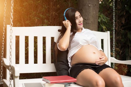 Asian young pregnant woman relaxing and listening music on swing in park garden with books. People and Entertainment concept. Happiness and pleasure concept. Single mom and Widow theme. 