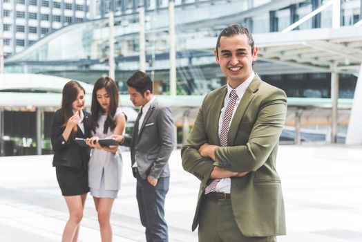 Smiling young businessman looking into camera with his arms crossed and teammates background in urban. Modern business office team at outdoors in city. Business people and Leadership lifestyle concept