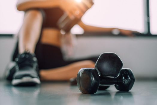 Close up of dumbbell with exercise woman lifestyle workout in gym fitness breaking relax after sport training with protein shake bottle background. Healthy lifestyle bodybuilding athlete muscles