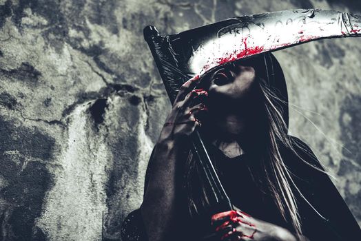 Witch licking blood on reaper. Female demon angel in black clothes and hood on grunge wall background. Halloween day and Mystery concept. Fantasy of magic theme. Afterlife and Death concept.