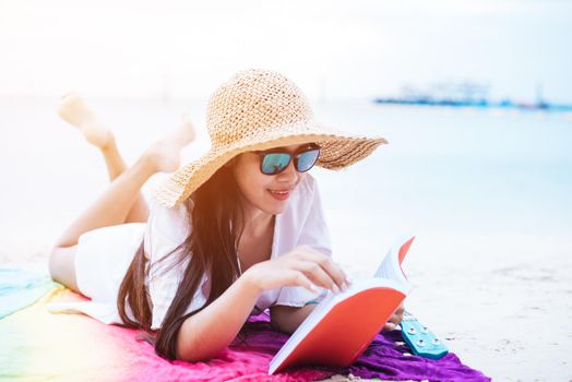 Beauty Asian woman have vacation on beach. Girl wearing wing hat and reading book on colorful mat near sea. Lifestyle and happy life concept. Travel and holiday theme.