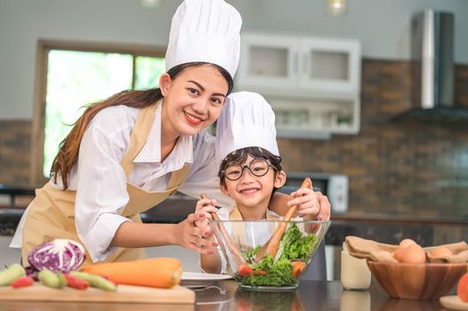 Beautiful Asian woman and cute little boy with eyeglasses prepare to cooking in kitchen at home. People lifestyles and Family. Homemade food and ingredients concept. Two Thai people looking at camera
