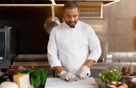 Handsome young African chef standing in professional kitchen in restaurant preparing a meal of meat and cheese vegetables. Portrait of man in cook uniform Cuts red pepper with a metal knife.