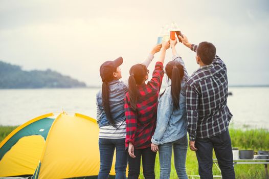 Back view of friendship clinking drinking bottle glass for celebrating in private party with mountain and lake view background. People lifestyle and travel on vacation concept. Picnic and camping tent