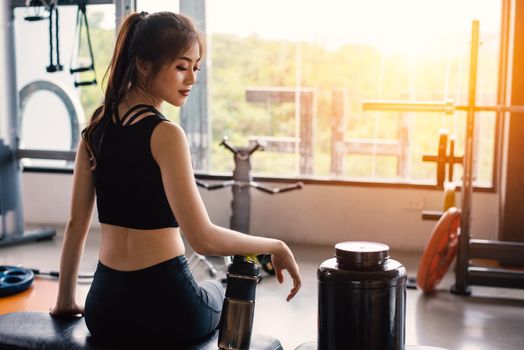 Sport woman sitting and resting after workout or exercise in fitness gym with protein shake or drinking water on floor. Relax concept. Strength training and Body build up theme. Warm and cool tone