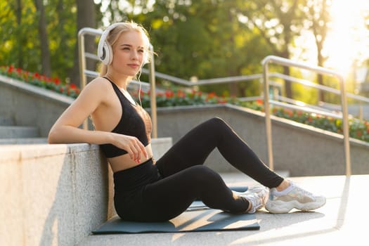 Sport and Fitness Young Adult Caucasian Woman Sitting Exercise Mat Concrete Floor Resting after morning Workout Outdoor Summer Park Athletic Female Take a Break listen Music Headphones