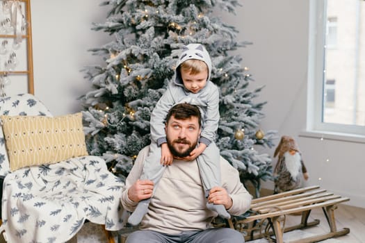 Holiday Christmas Handsome father playing with small cute little boy near decorated New year tree at home Family tradition positive son sitting on dad shoulders