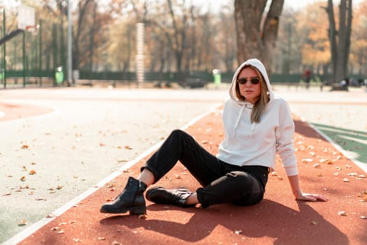 Outdoor portrait of young beautiful woman with long in sunglasses and a white hooded sweater sitting on the sportsground track. youth culture summer pastime