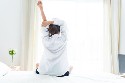 Back view of woman stretching in morning after waking up on bed near window. Holiday and Relax concept. Lazy day and Working day concept. Office woman and worker in daily life theme